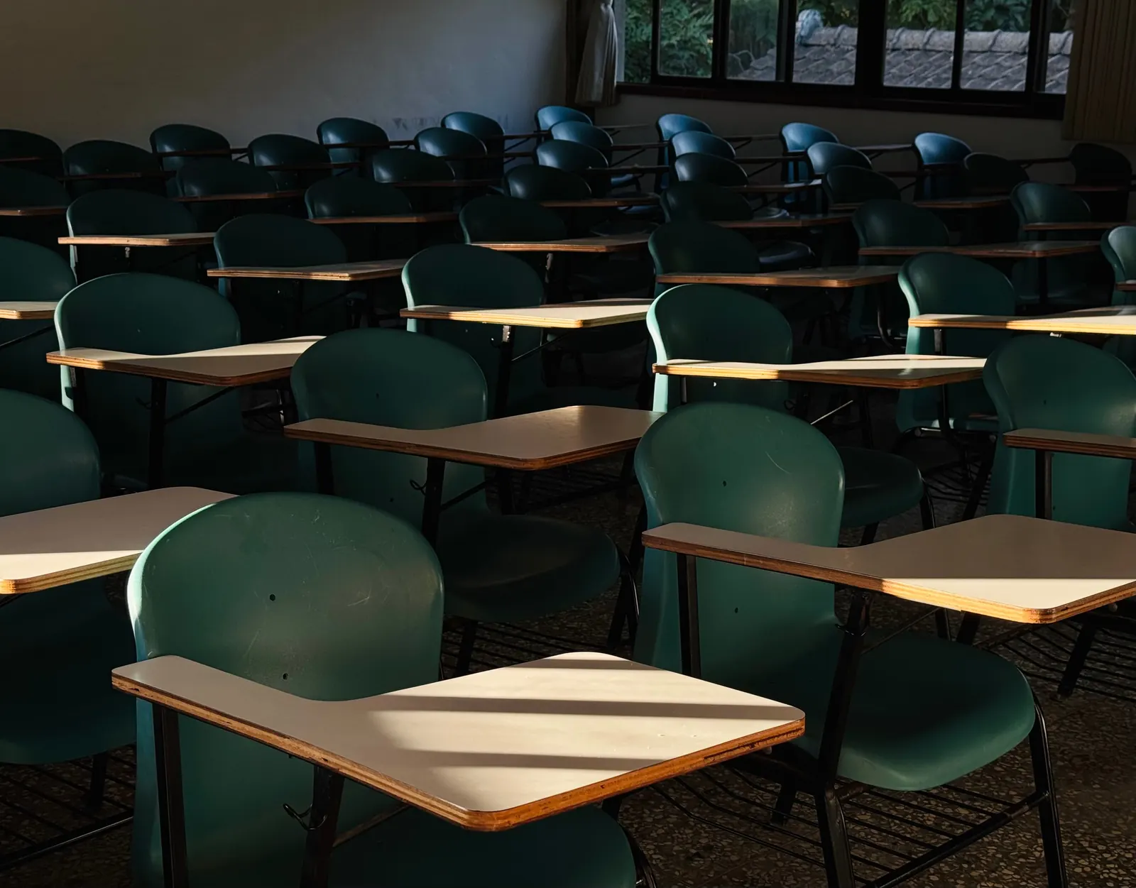 Rows of green classroom chairs with pale writing tablets, striped by afternoon sun from a window