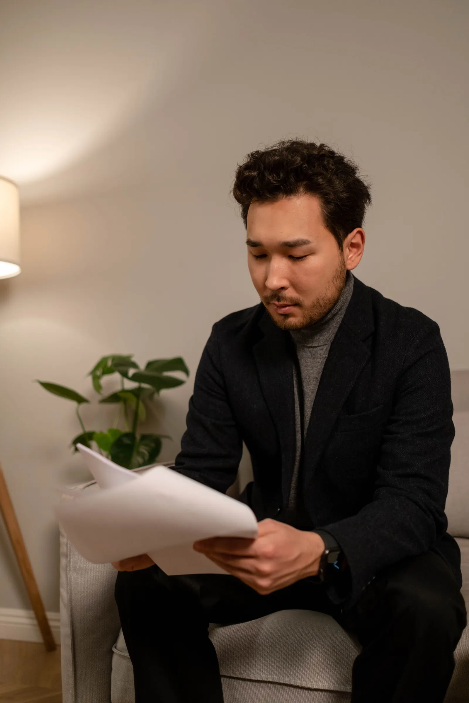 A man in a dark jacket sits in a quiet room reading through printed pages he holds in both hands
