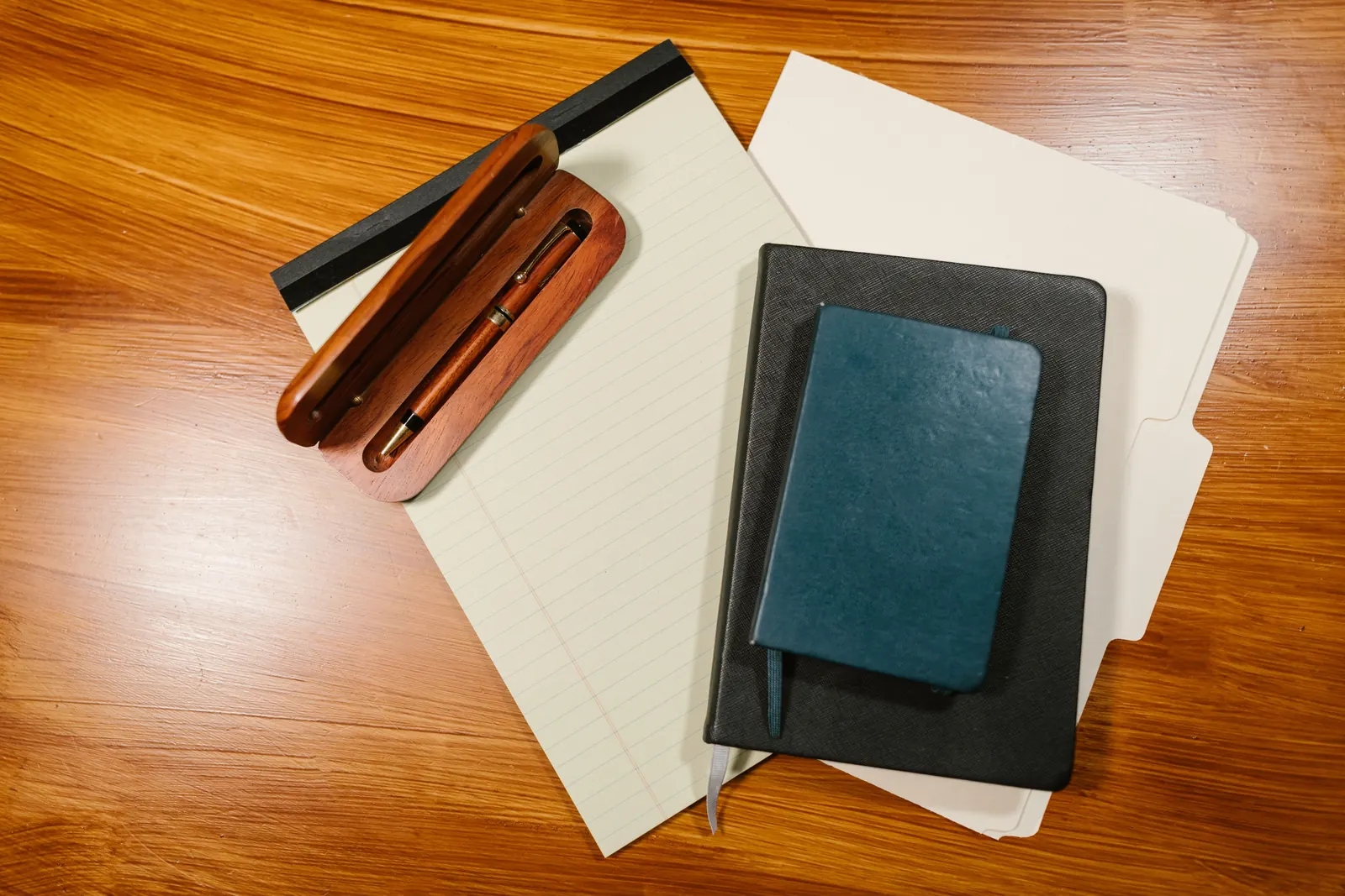 A dark leather folder, a blue notebook and a lined pad with a wooden pen case on a warm wooden desk