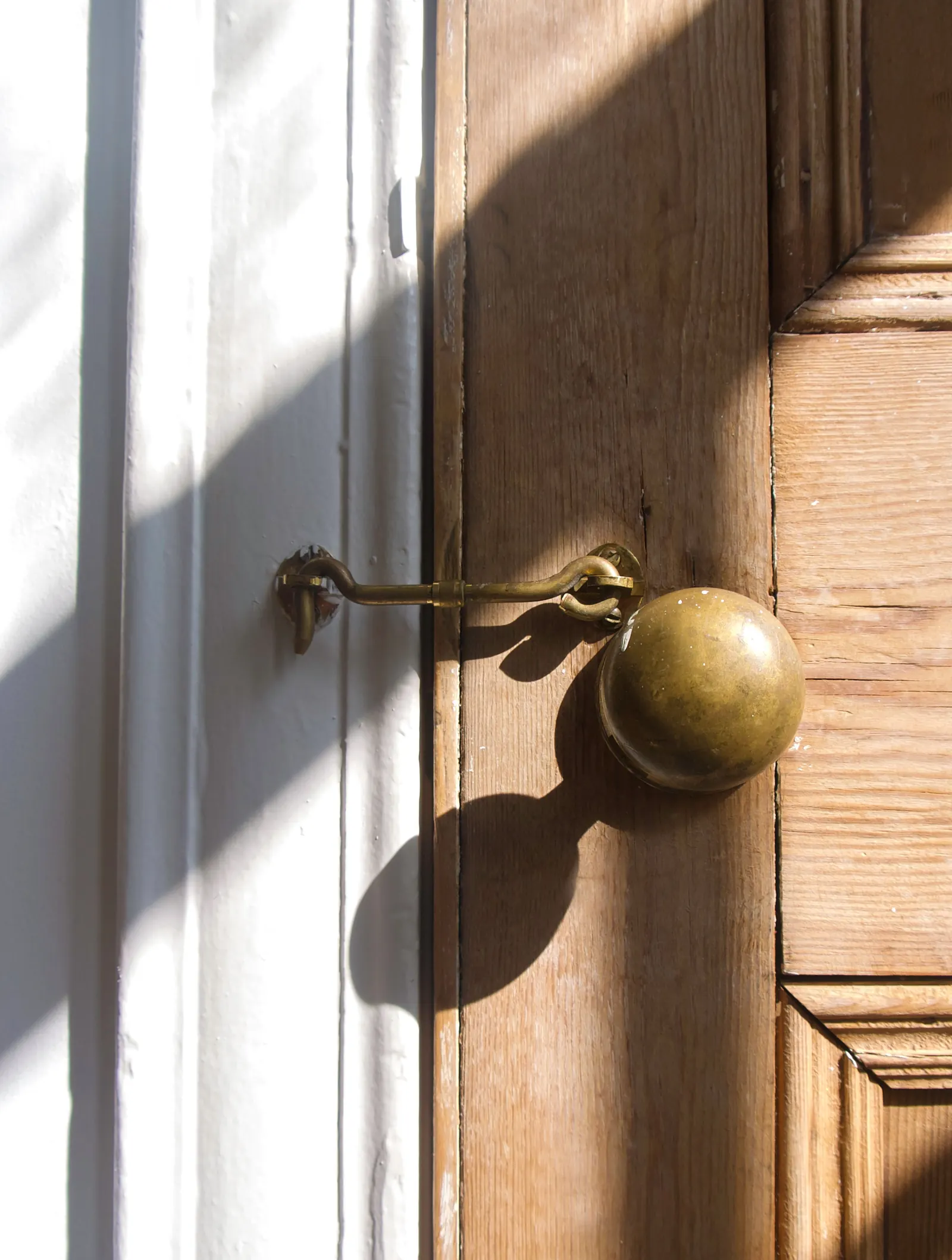 A brass hook latch fastens a sunlit wooden door beside a round brass knob