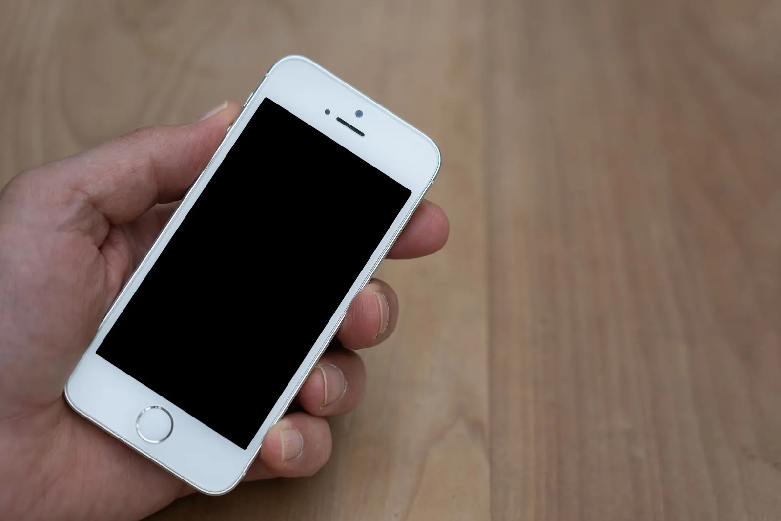 A man's hand holds a white smartphone with a dark blank screen over a wooden table