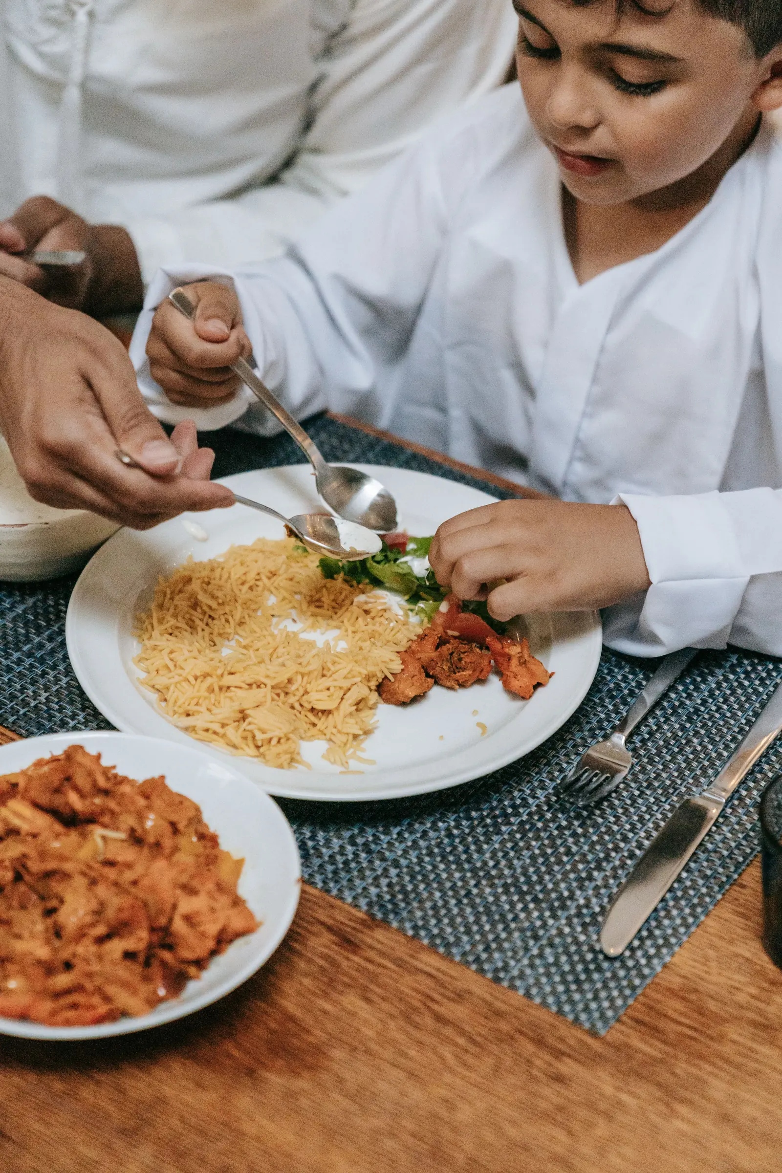 A boy in a white kurta eats rice and chicken at a wooden table as a man's hand passes a spoonful