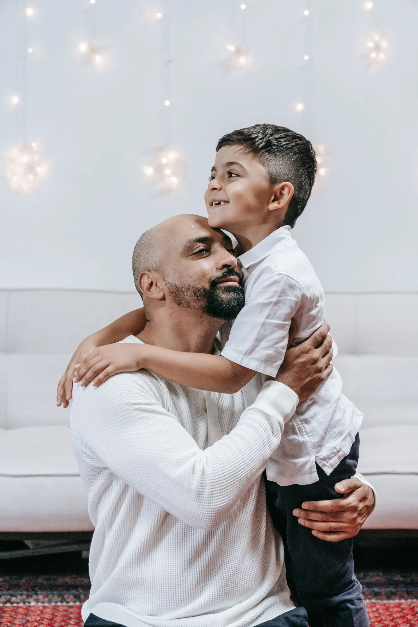 A father kneels to hug his young son in a white shirt goodbye, the boy smiling over his shoulder