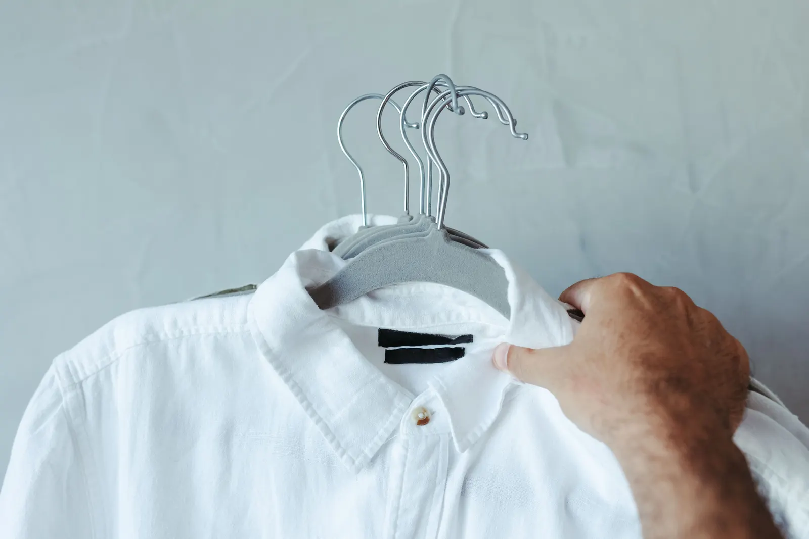 A man's hand holds a crisp white collared shirt on a grey hanger against a pale blue-grey wall