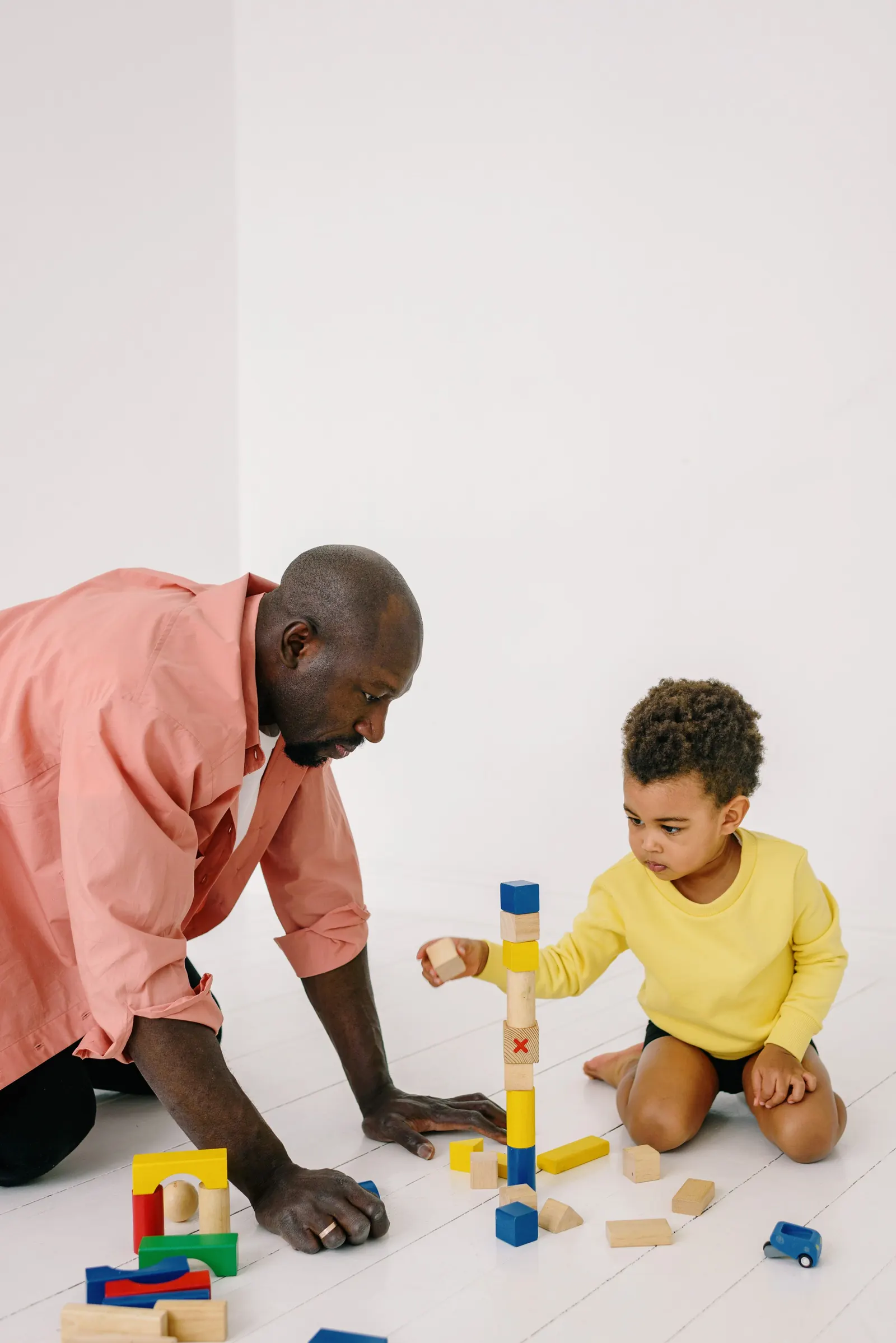 A man kneels on the floor beside a toddler boy building a tower of wooden blocks