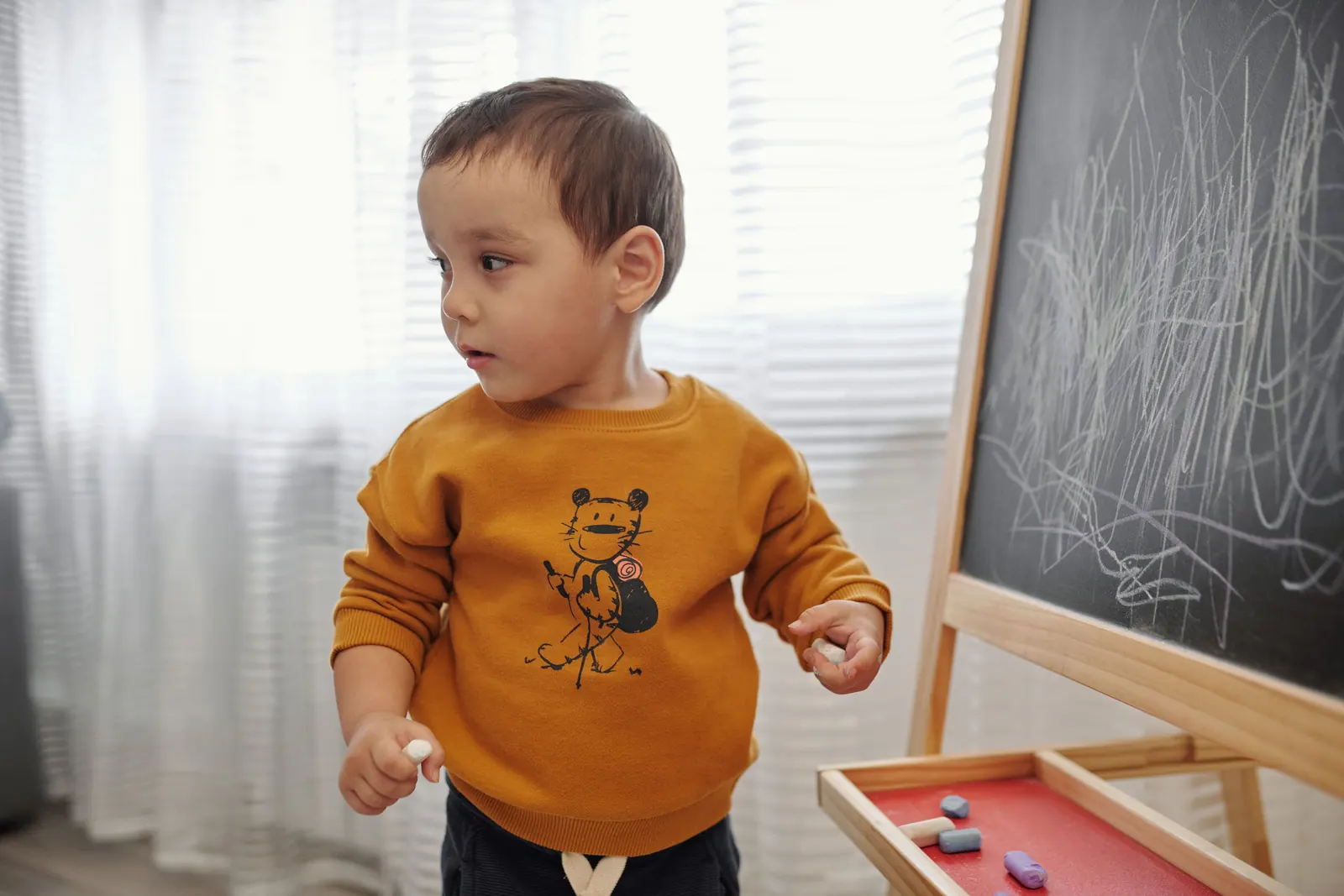 A toddler boy in a mustard sweatshirt holds chalk beside a chalkboard easel