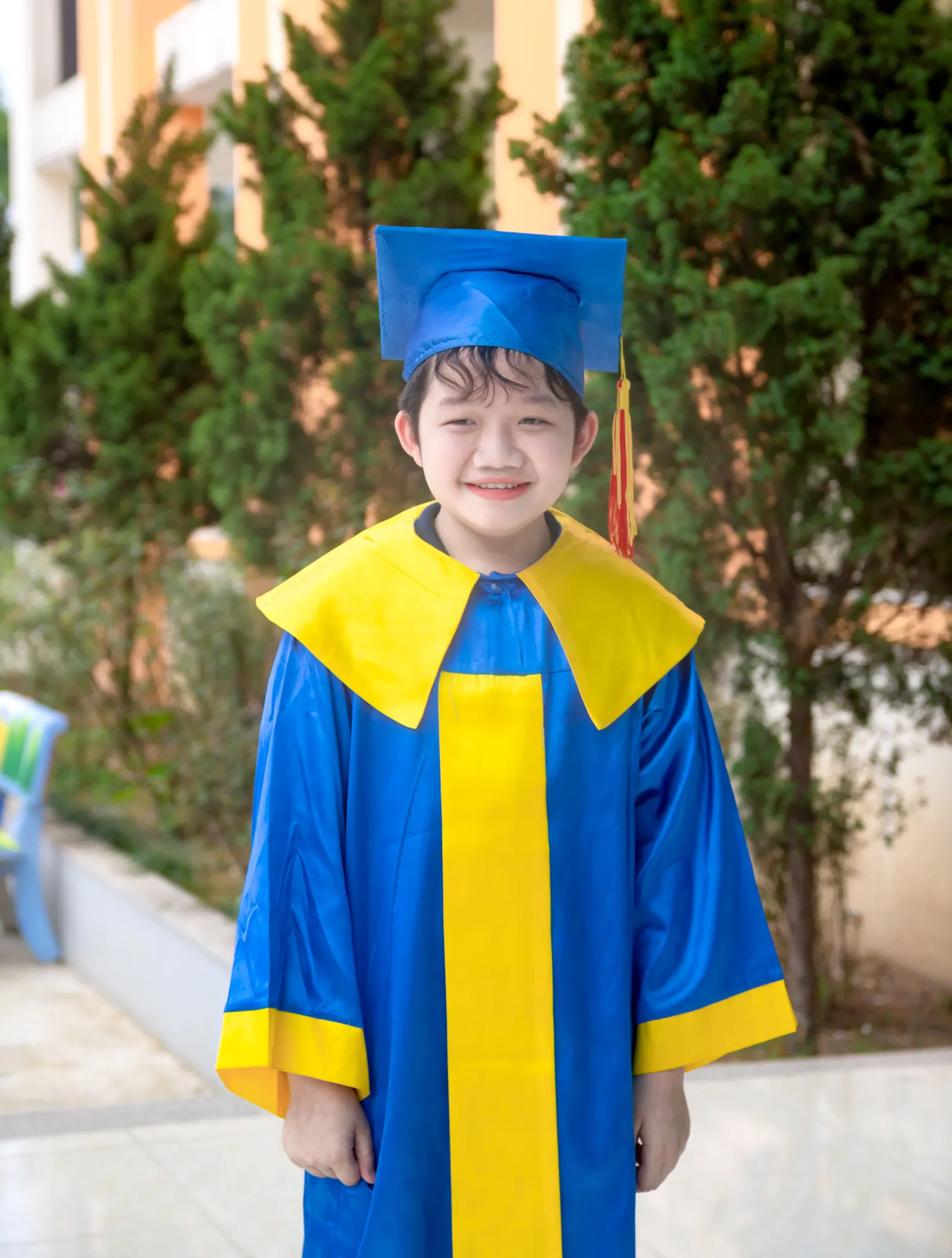 A smiling small boy in a blue and yellow graduation cap and gown stands outdoors