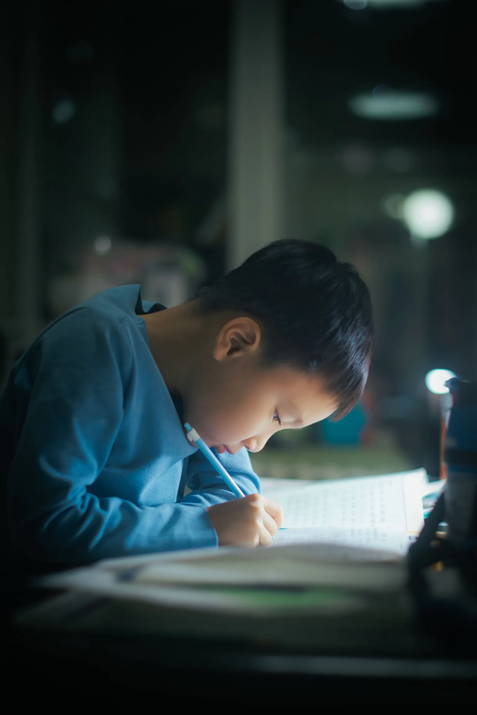 A boy in a blue shirt bends over his desk writing carefully under a lamp
