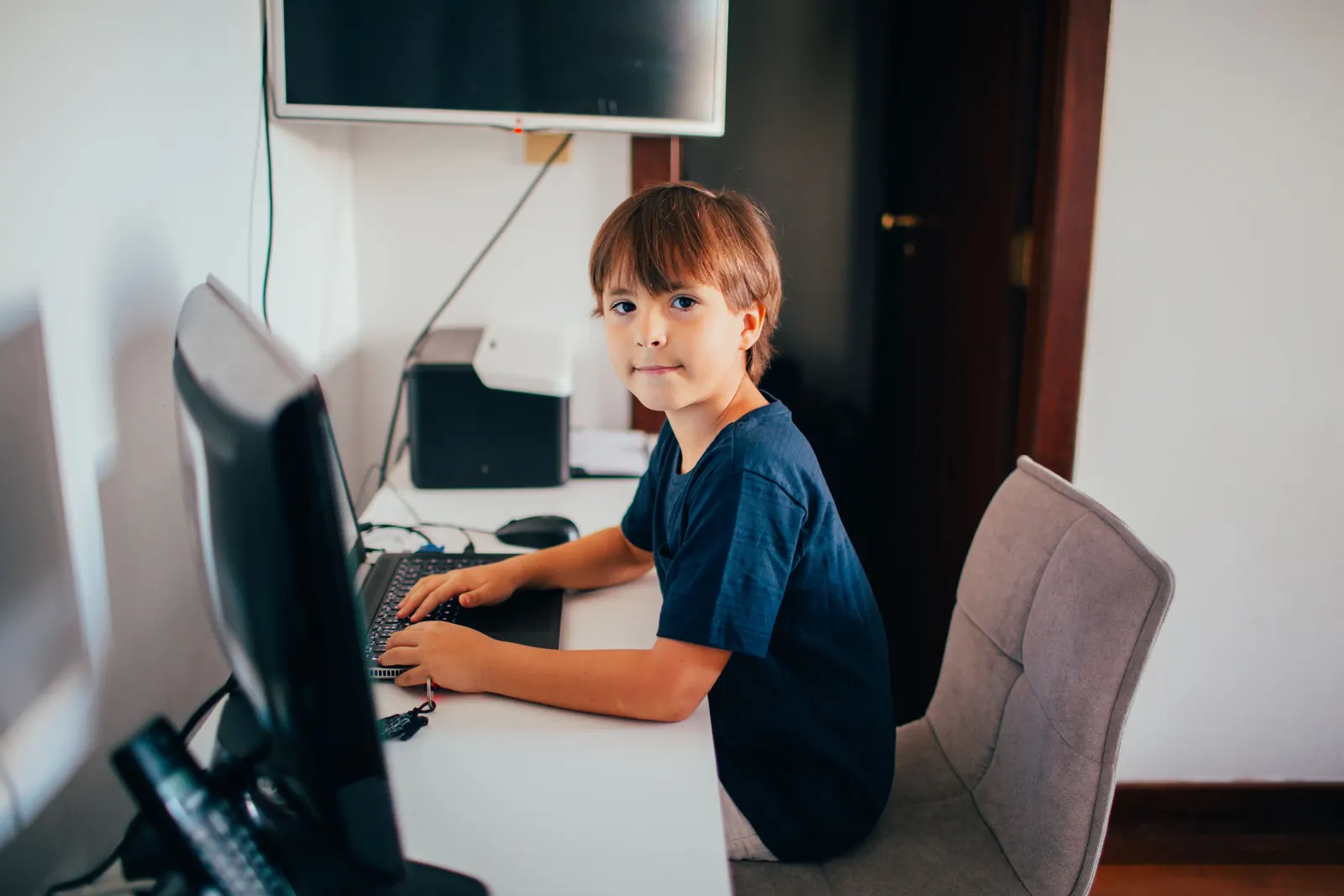 A young boy types at a desktop computer and looks round at the camera