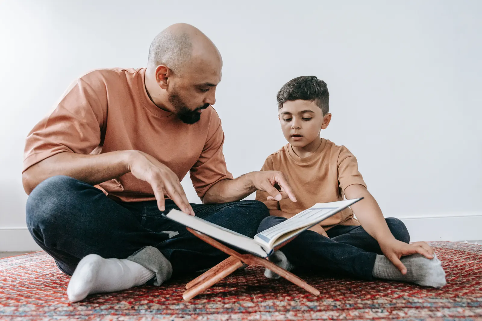 A bearded man sitting cross-legged on a rug points to the Qur'an on a wooden stand as a young boy reads