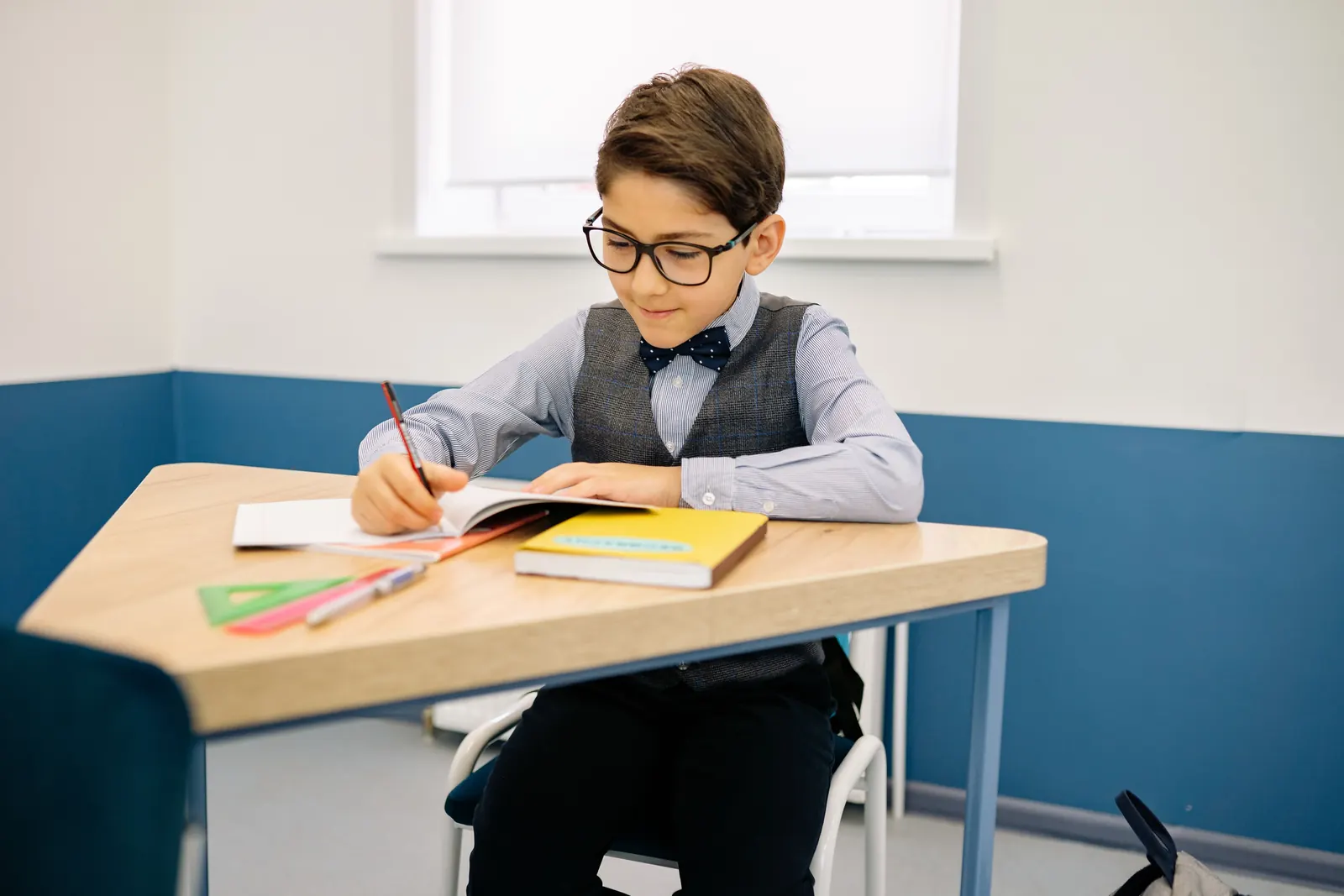 A boy in glasses and a bow tie writes in his notebook at a classroom desk, rulers and a set square beside him