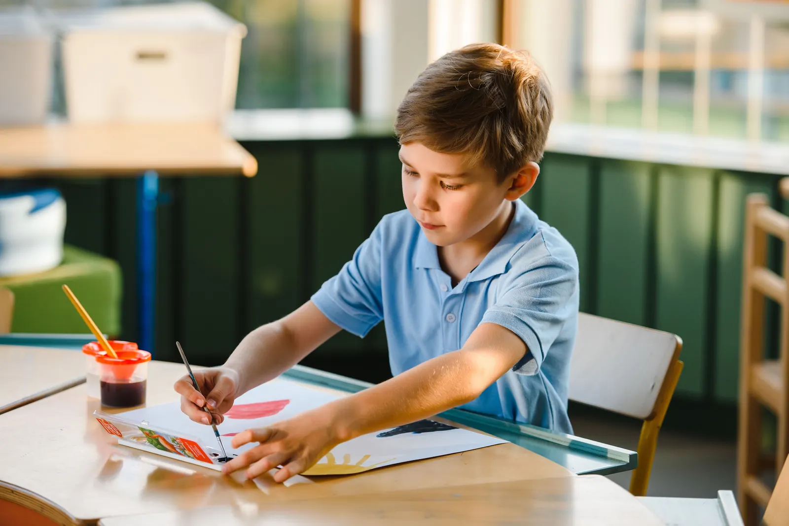 A boy in a blue polo paints with a brush at a sunlit classroom table