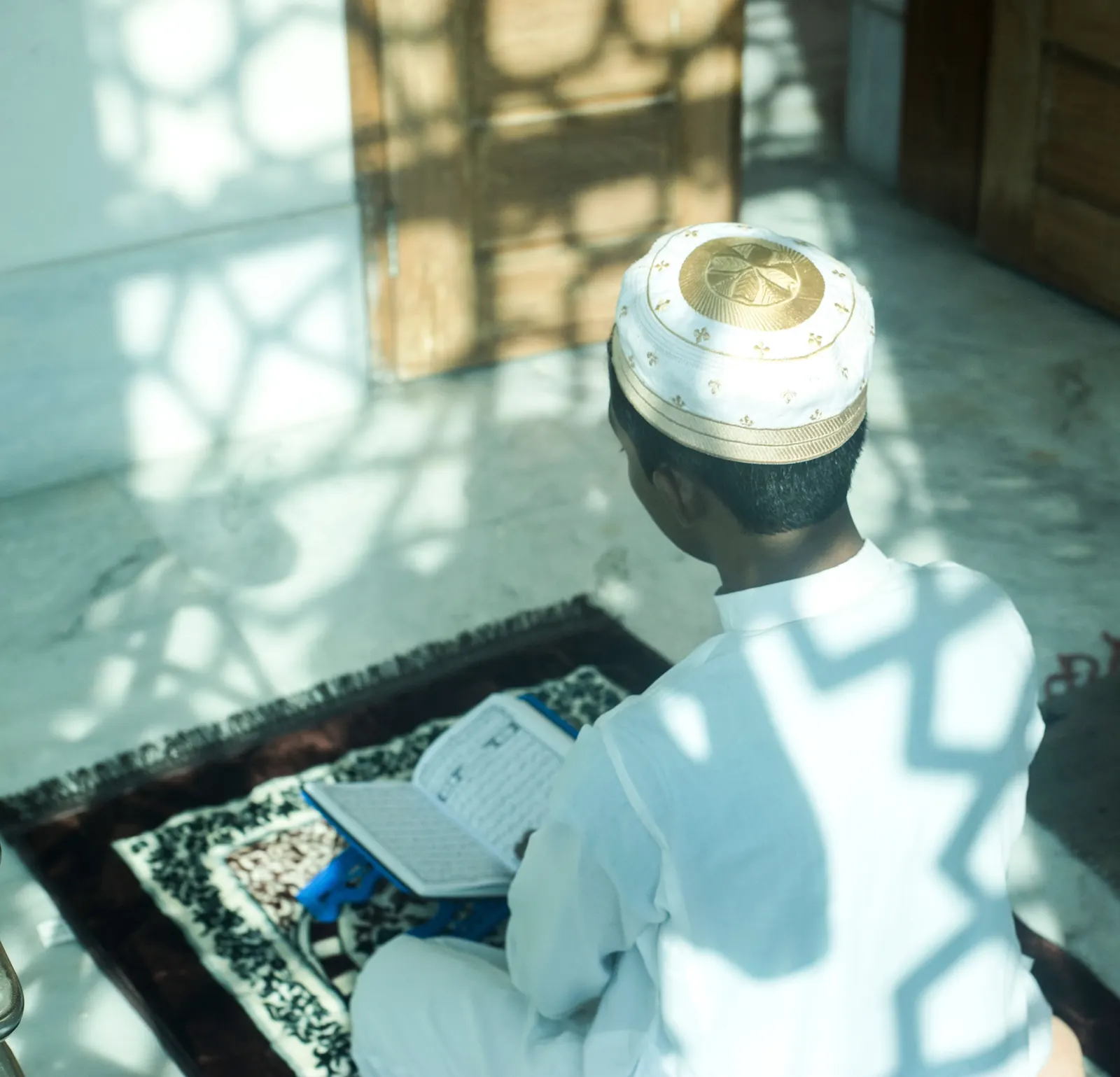 A boy in a white-and-gold kufi reads a Qur'an on a prayer mat, lattice window light falling across him