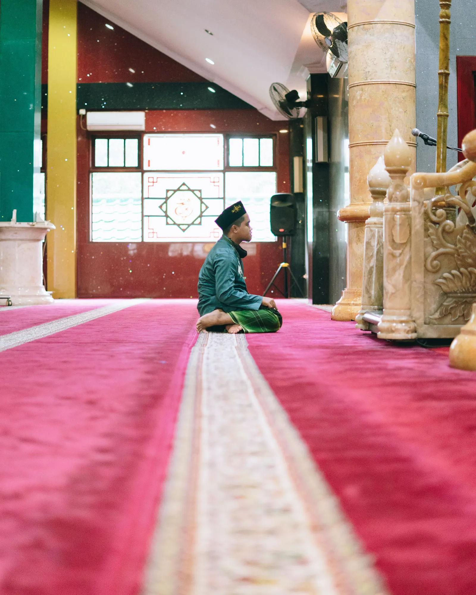 A boy in a dark cap and teal shirt sits on a crimson mosque carpet facing the carved minbar and microphone