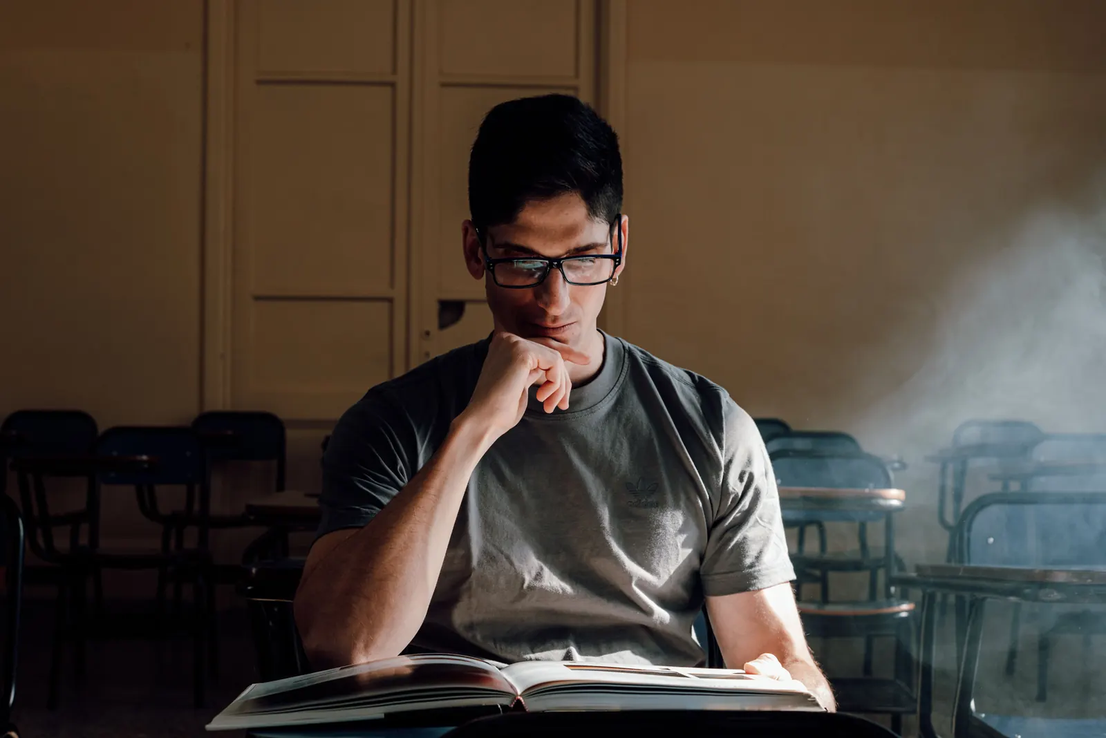 A young man in glasses reads a large book in a quiet, sunlit classroom