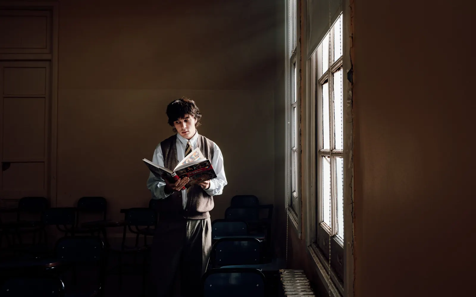A teenage boy stands reading a book by a tall window in a dim classroom