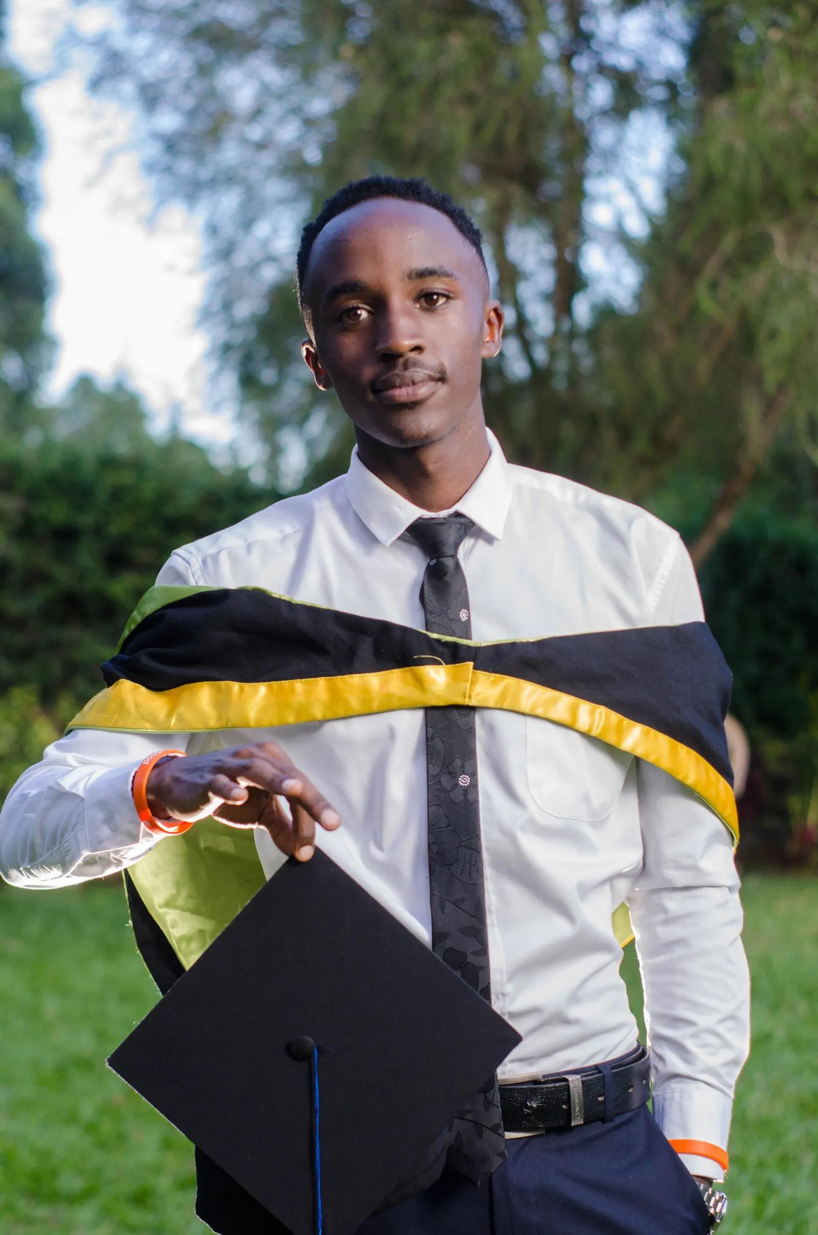 A young man in a white shirt, tie and graduation stole holds his mortarboard outdoors