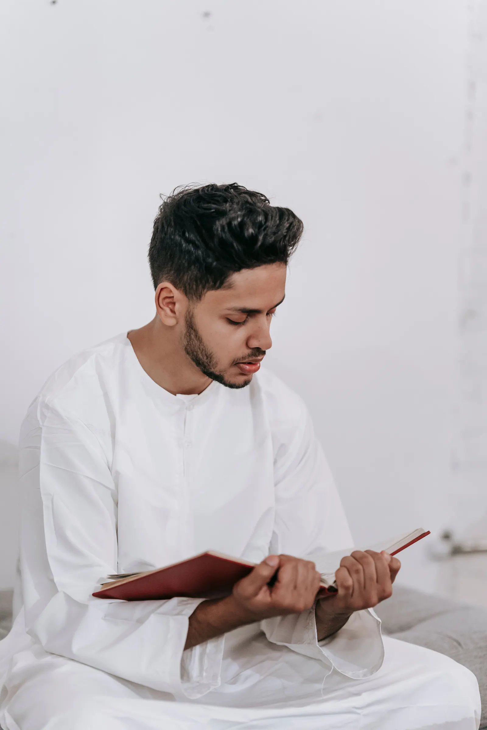 A young man in a white thobe sits reading a red-covered book