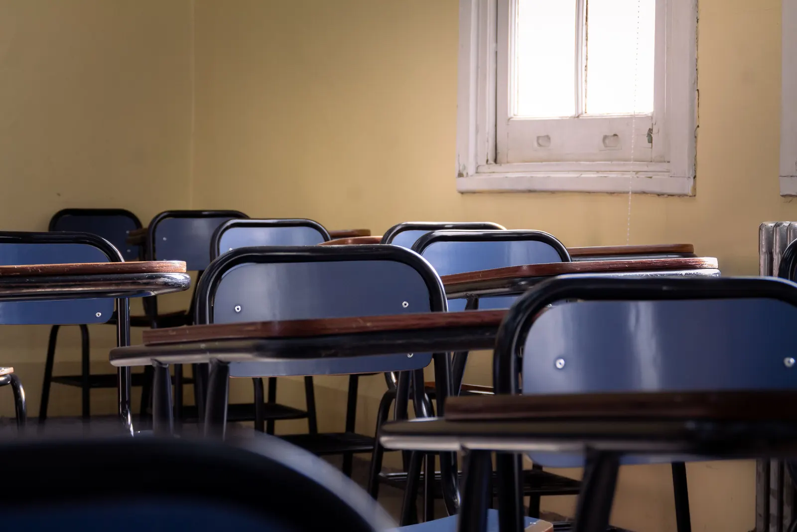 Rows of empty blue chairs and wooden desks in a warm classroom with a tall window