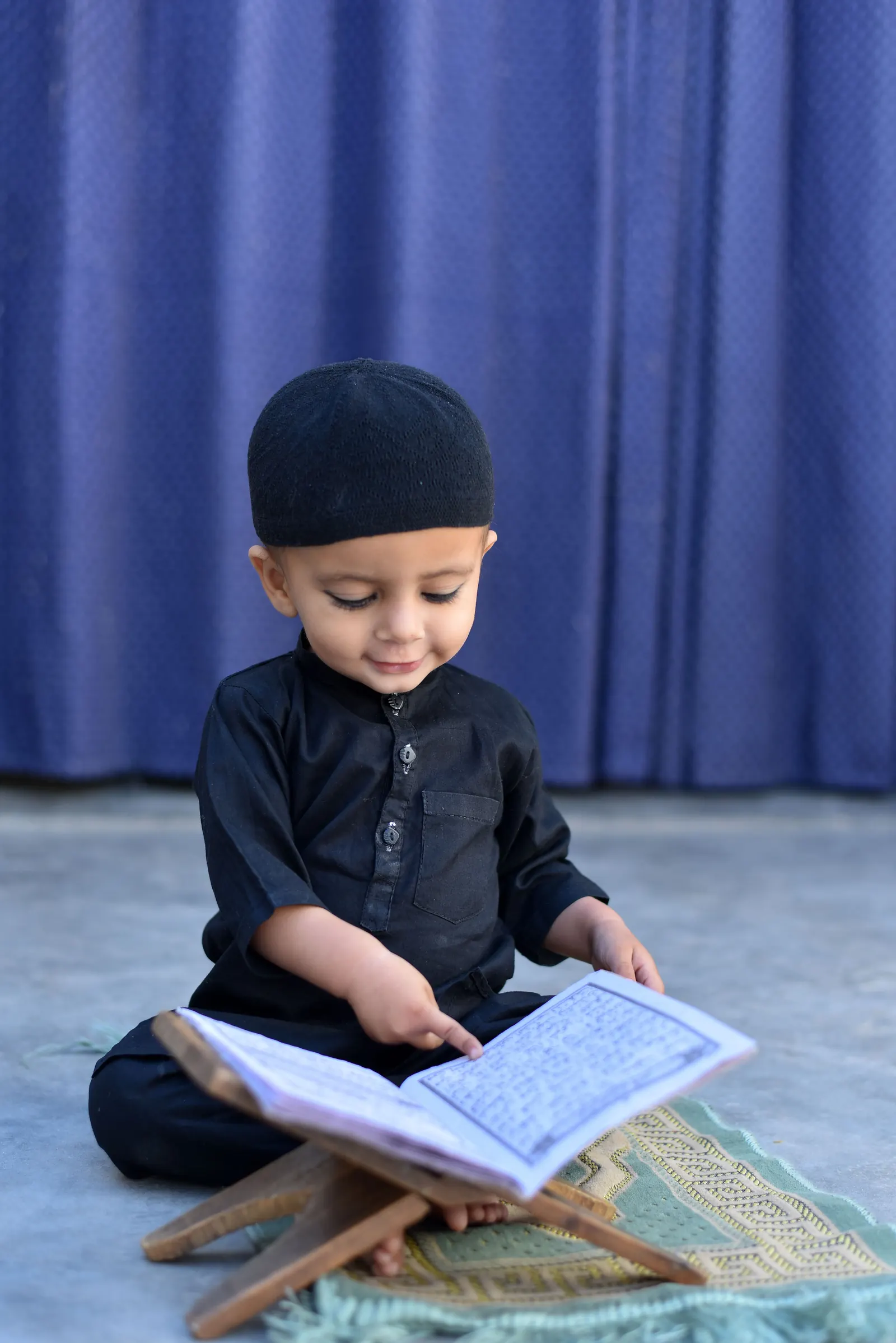 A little boy in a black kufi and kurta traces the lines of a mushaf on a wooden rehal