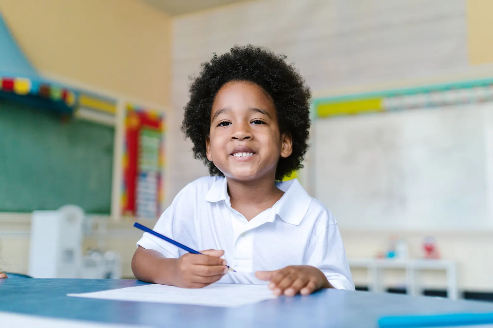 A smiling young boy in a white polo shirt holds a pencil over his worksheet at a classroom table