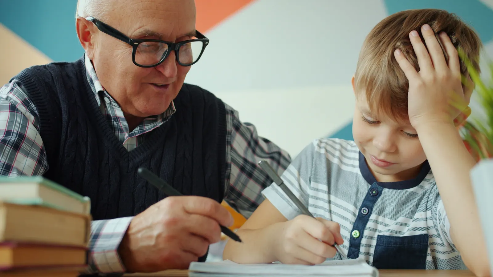 A gray-haired man in glasses sits beside a young boy and guides him as the boy writes in a notebook