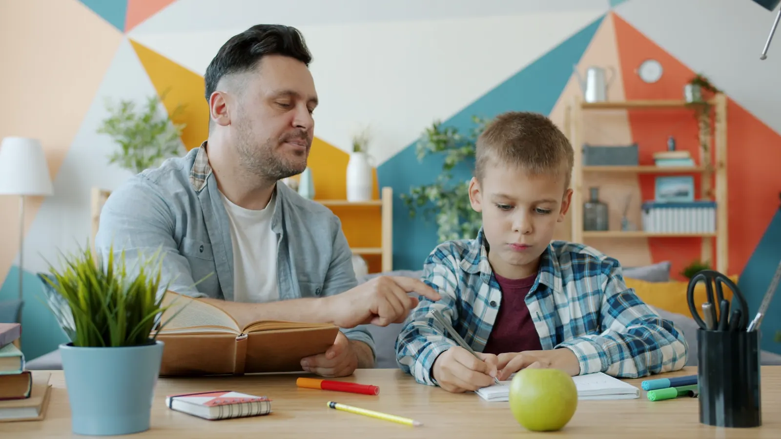A man points to the page as a young boy in a plaid shirt writes at a table