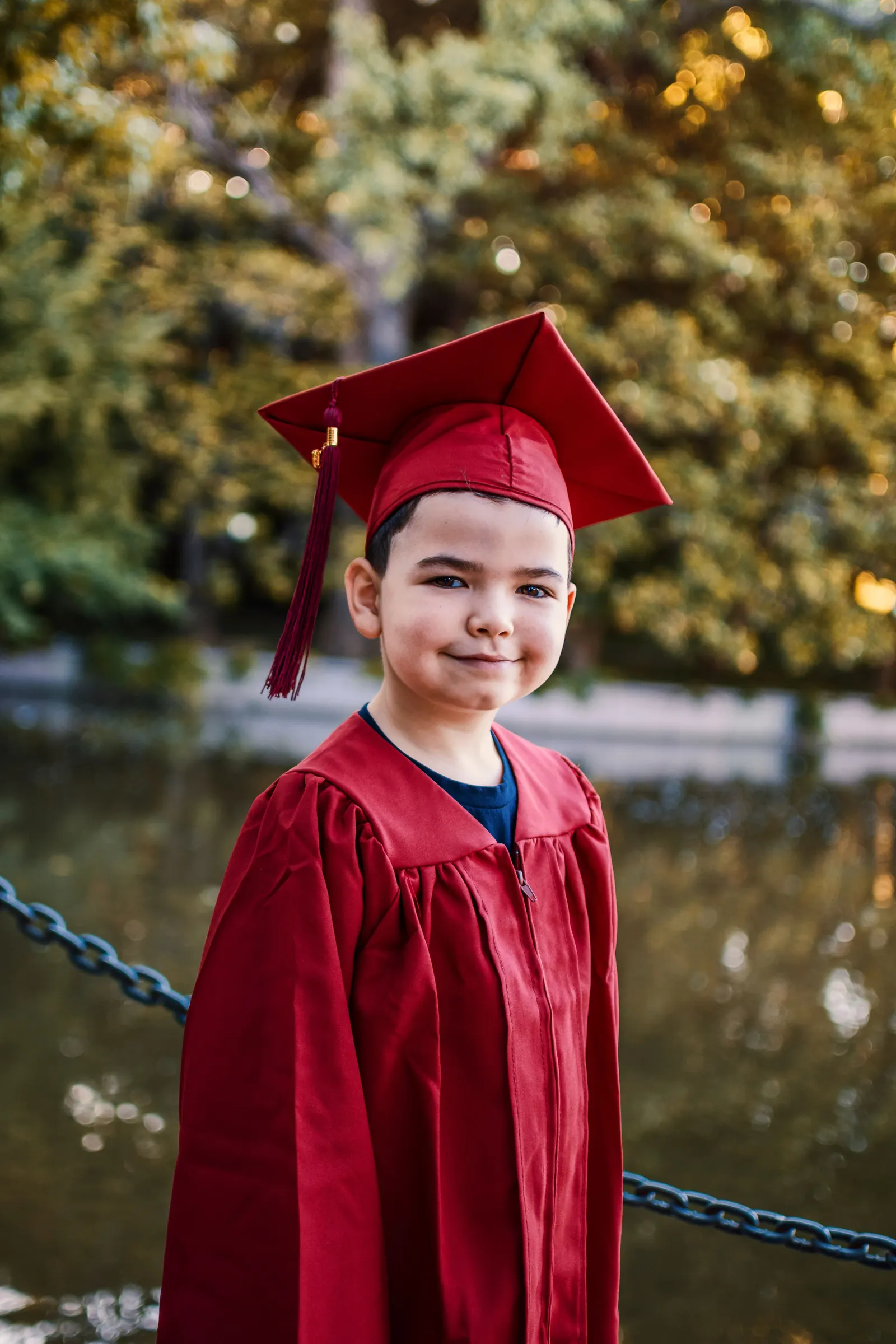A young boy in a red graduation cap and gown smiles beside a pond under autumn trees