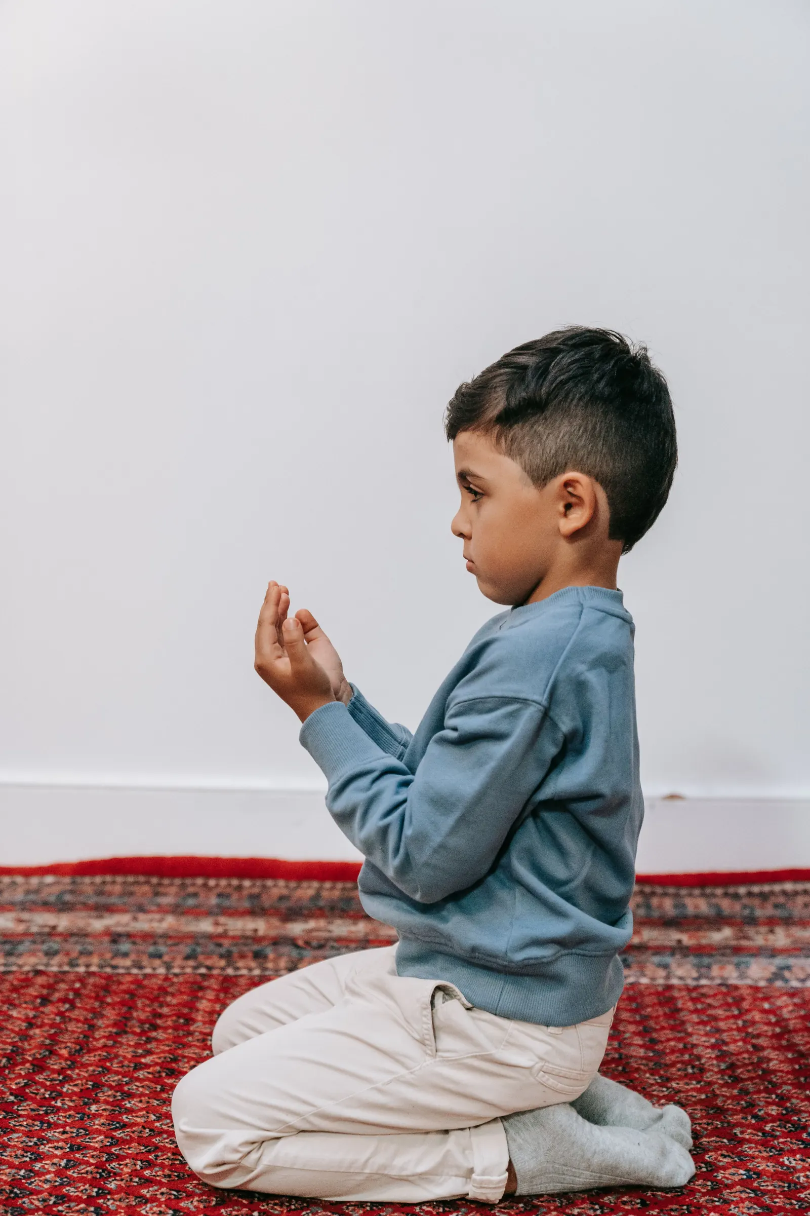 A young boy kneels on a red prayer rug with his hands raised in dua