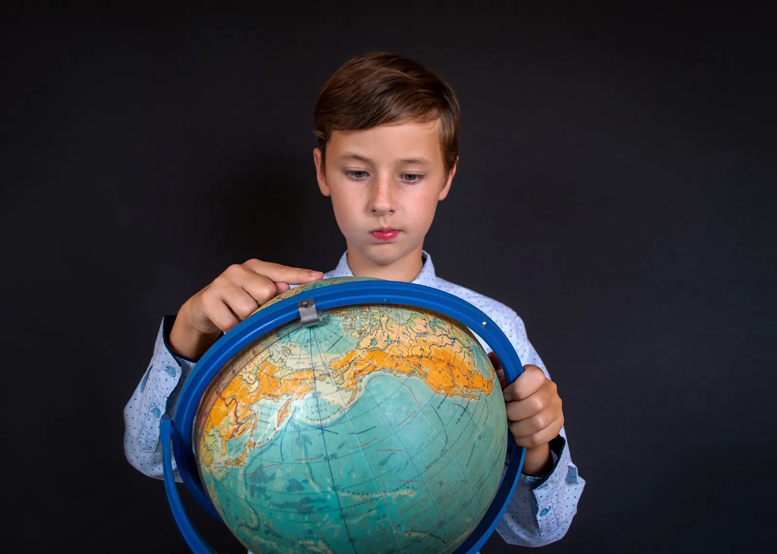 A boy in a patterned shirt holds a large desk globe against a dark background