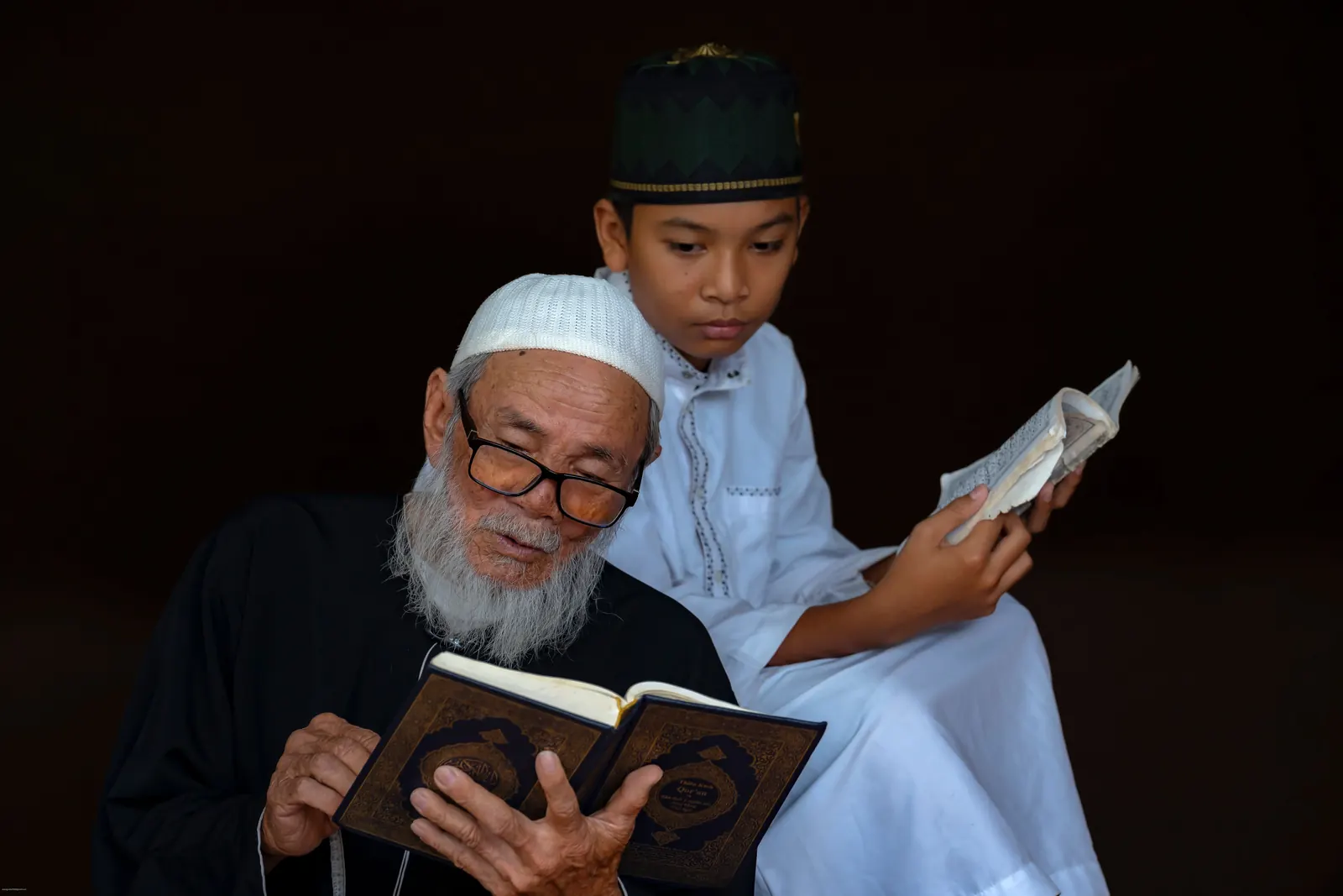 An elderly bearded man in a white cap reads the Qur'an while a boy in a kufi reads beside him