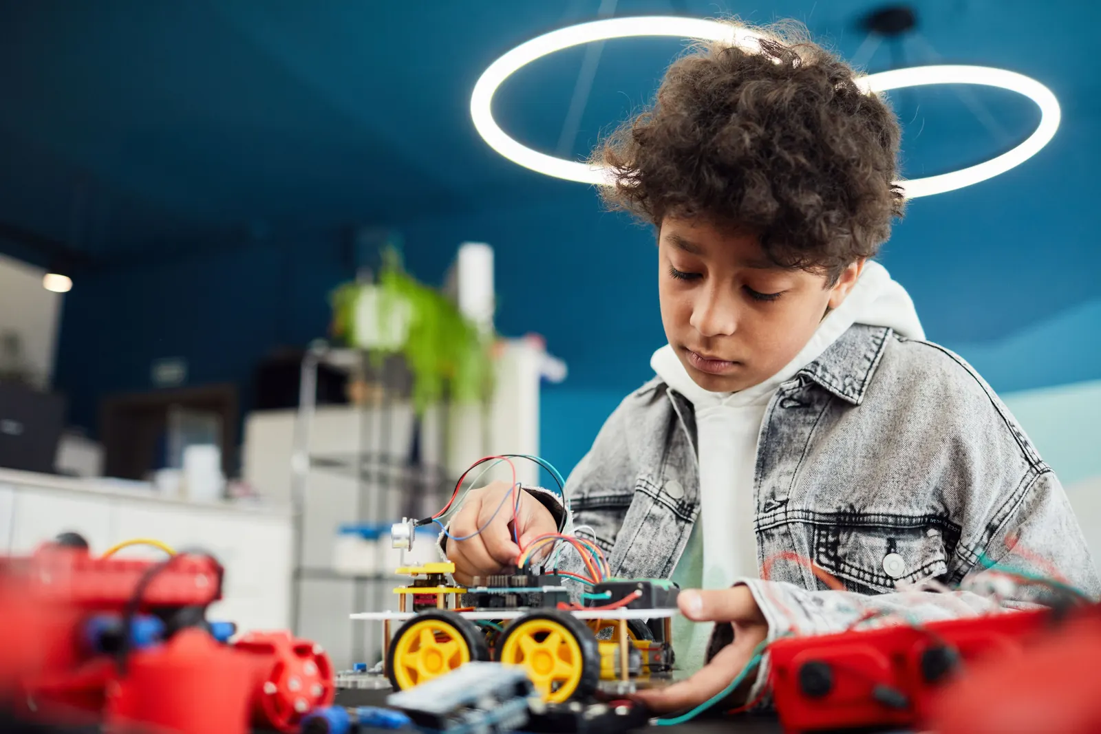 A boy in a denim jacket wires up a small wheeled robot on a workbench scattered with parts