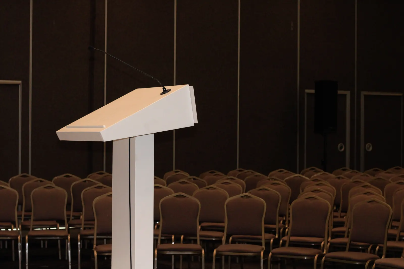 A white speaker's lectern with a microphone before rows of empty chairs in a dark hall