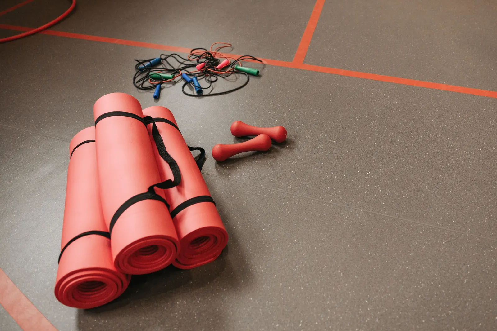 Rolled red exercise mats, jump ropes and small dumbbells laid out on a gym floor