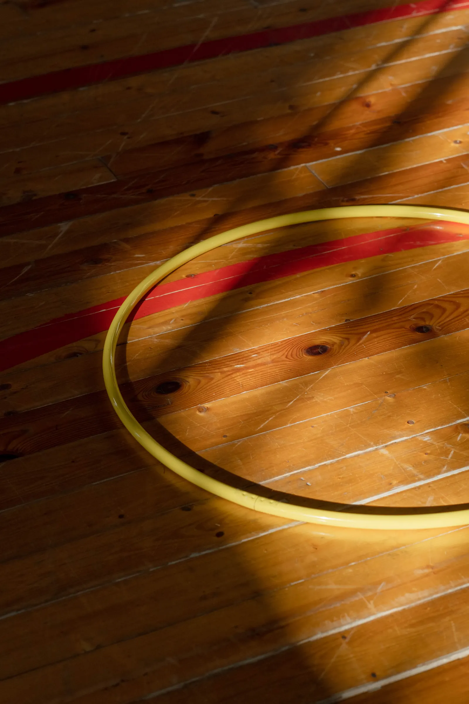 A yellow hula hoop lying on a wooden gym floor striped with afternoon sunlight