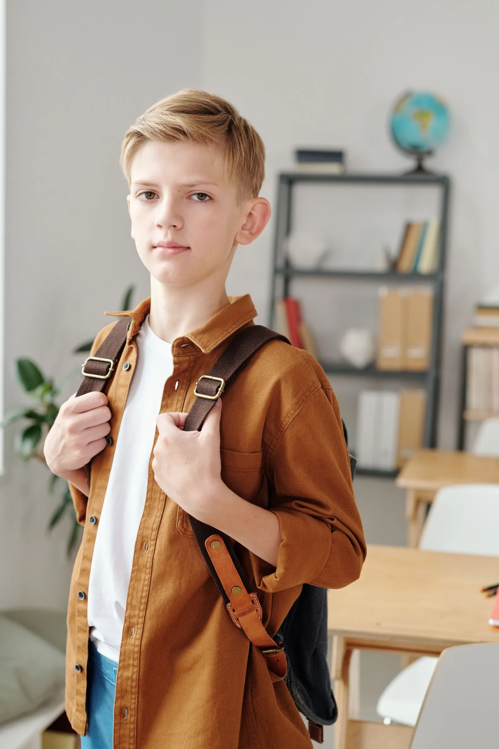 A boy in a tan shirt holds the straps of his backpack in a light classroom with shelves and a globe