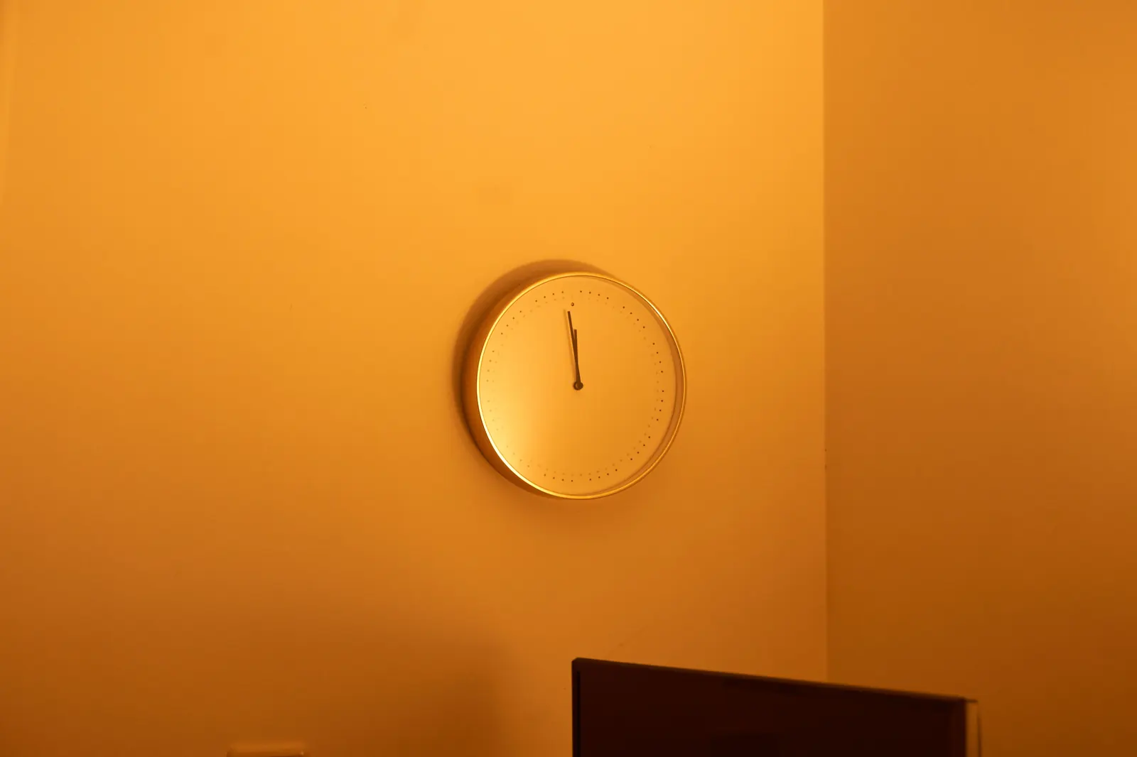 A plain round wall clock with a slim gold rim on a warm orange wall, its hands near twelve