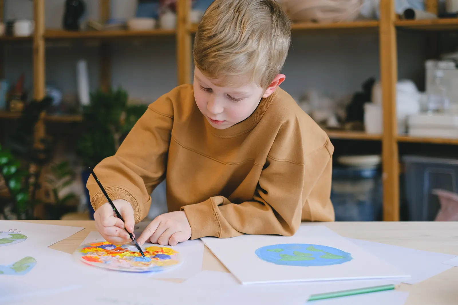 A young boy in a mustard sweatshirt paints a colourful circle with a fine brush at a table covered in paper