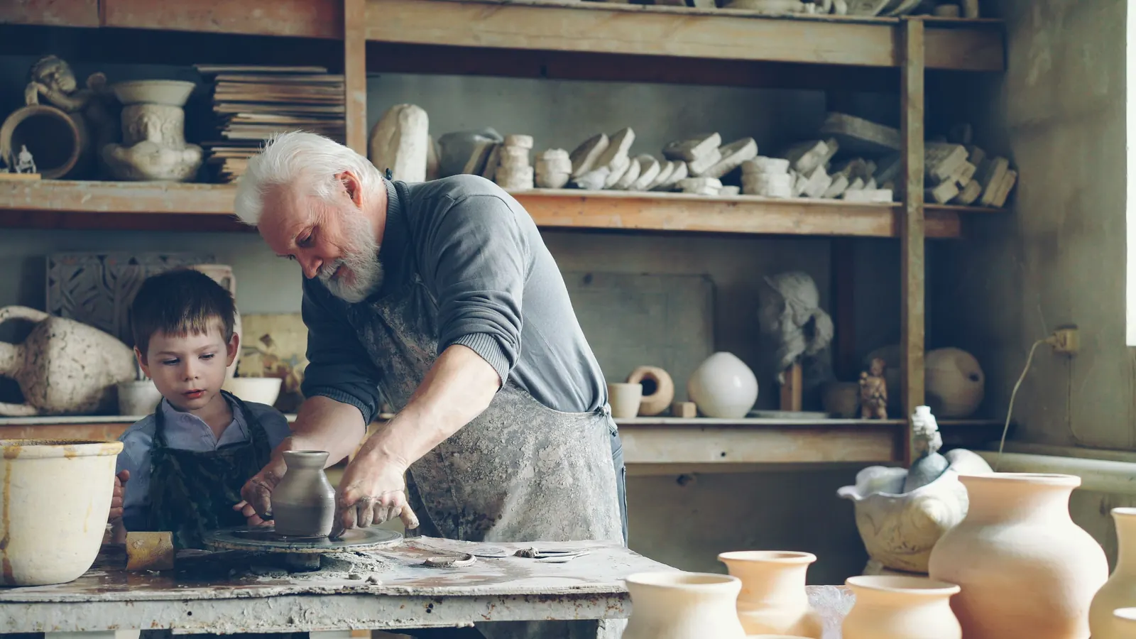 A white-haired man guides a young boy at the potter's wheel in a workshop lined with clay pots
