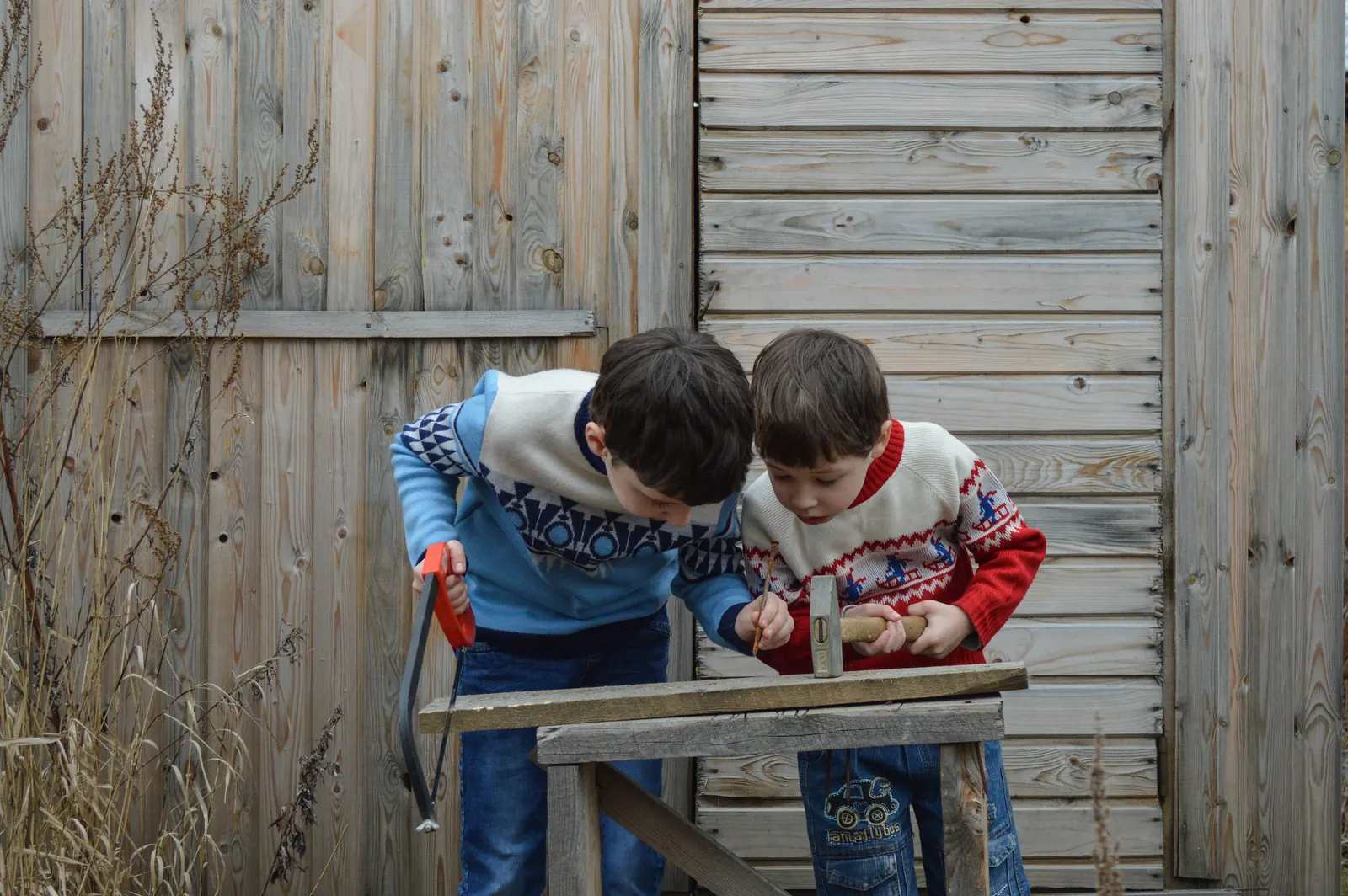 Two brothers in patterned sweaters lean over a sawhorse together, one with a saw and one with a hammer