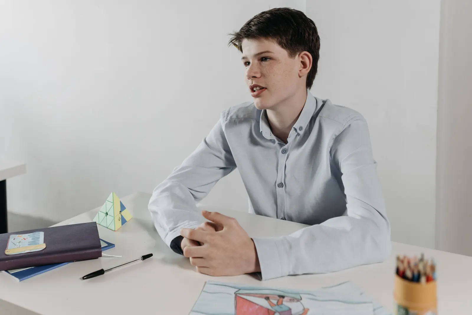 A teenage boy in a light blue shirt sits at a white table, hands clasped, speaking up as if leading a meeting