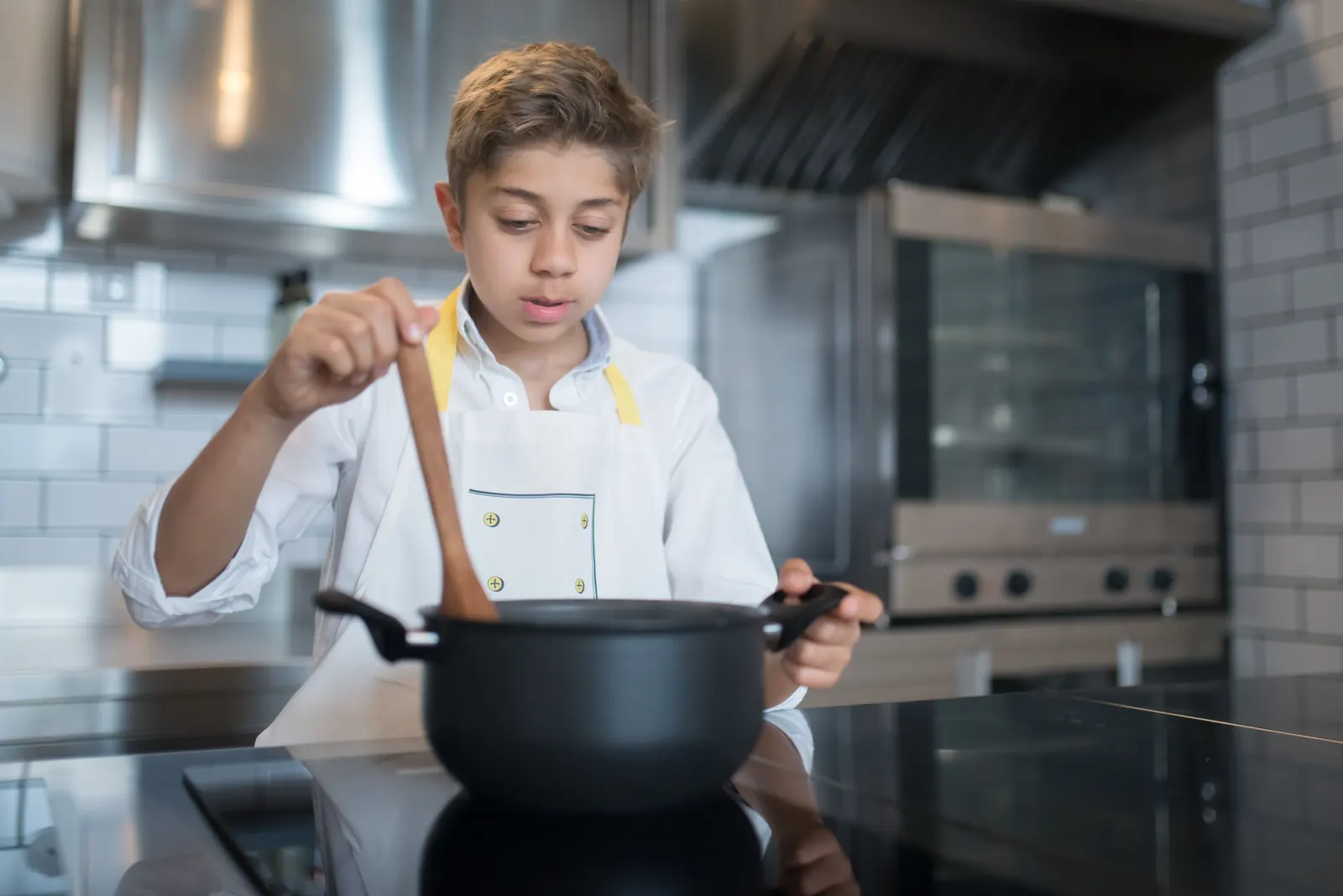 A boy in a white apron stirs a pot with a wooden spoon at a kitchen hob