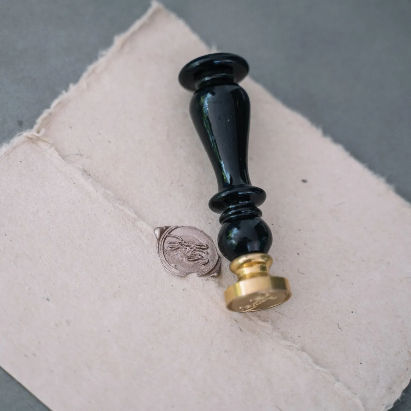 A black and brass wax-seal stamp lying on deckle-edged handmade paper beside a pressed silver wax seal