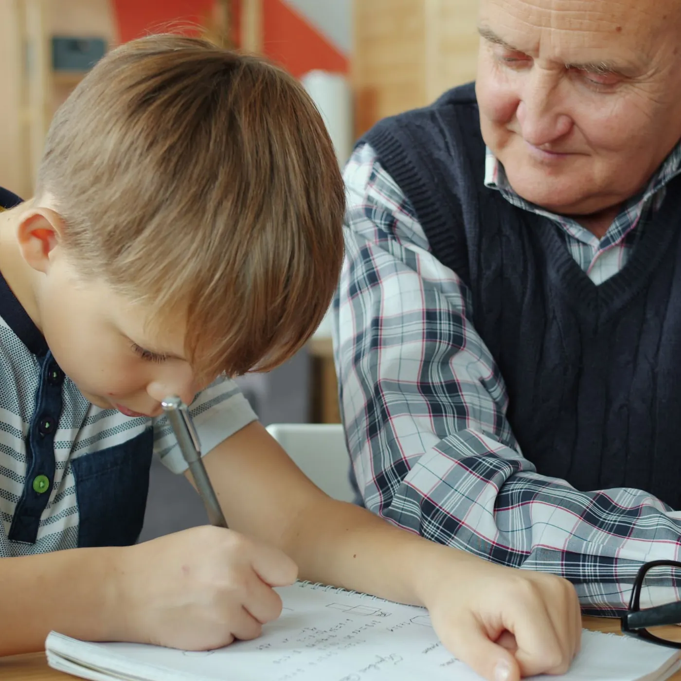 An older man in a sweater vest smiles as he watches a young boy write carefully in a spiral notebook at a wooden table