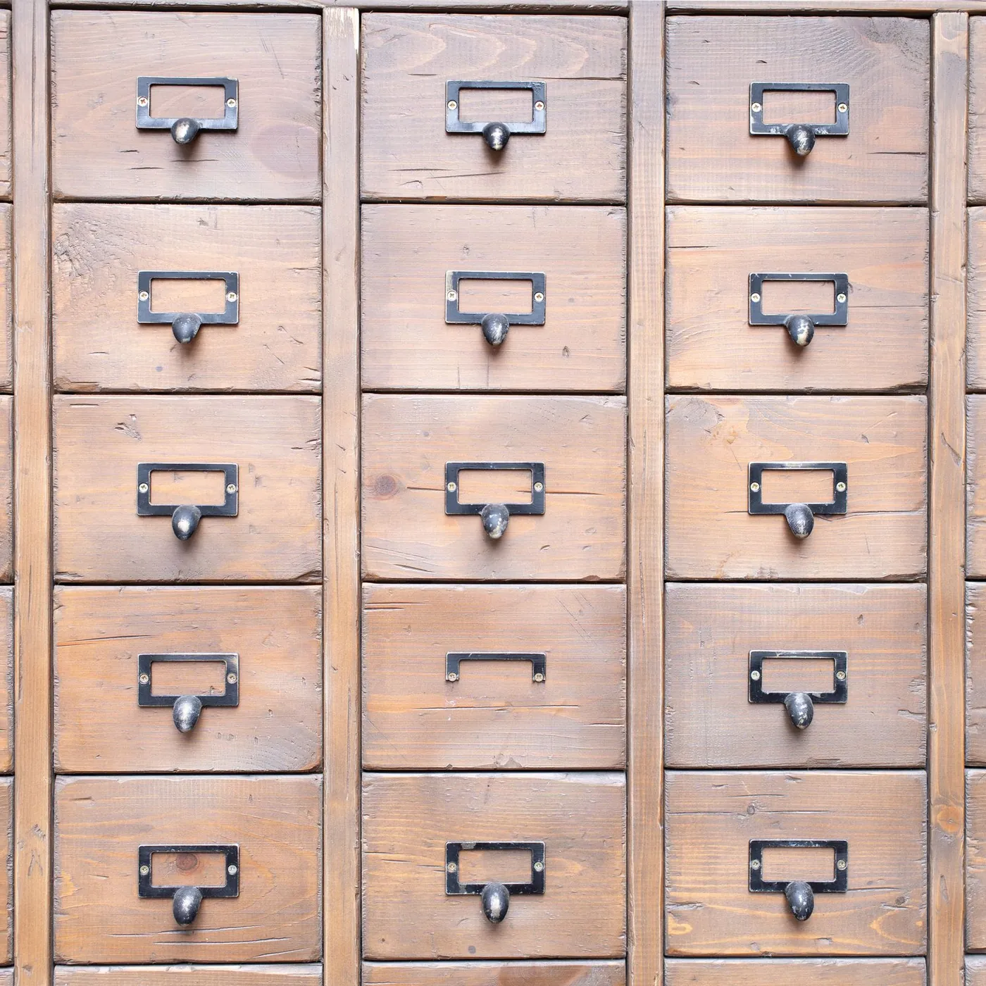 A wall of worn pine drawers in neat rows, each with a dark metal pull and an empty label holder, in warm light