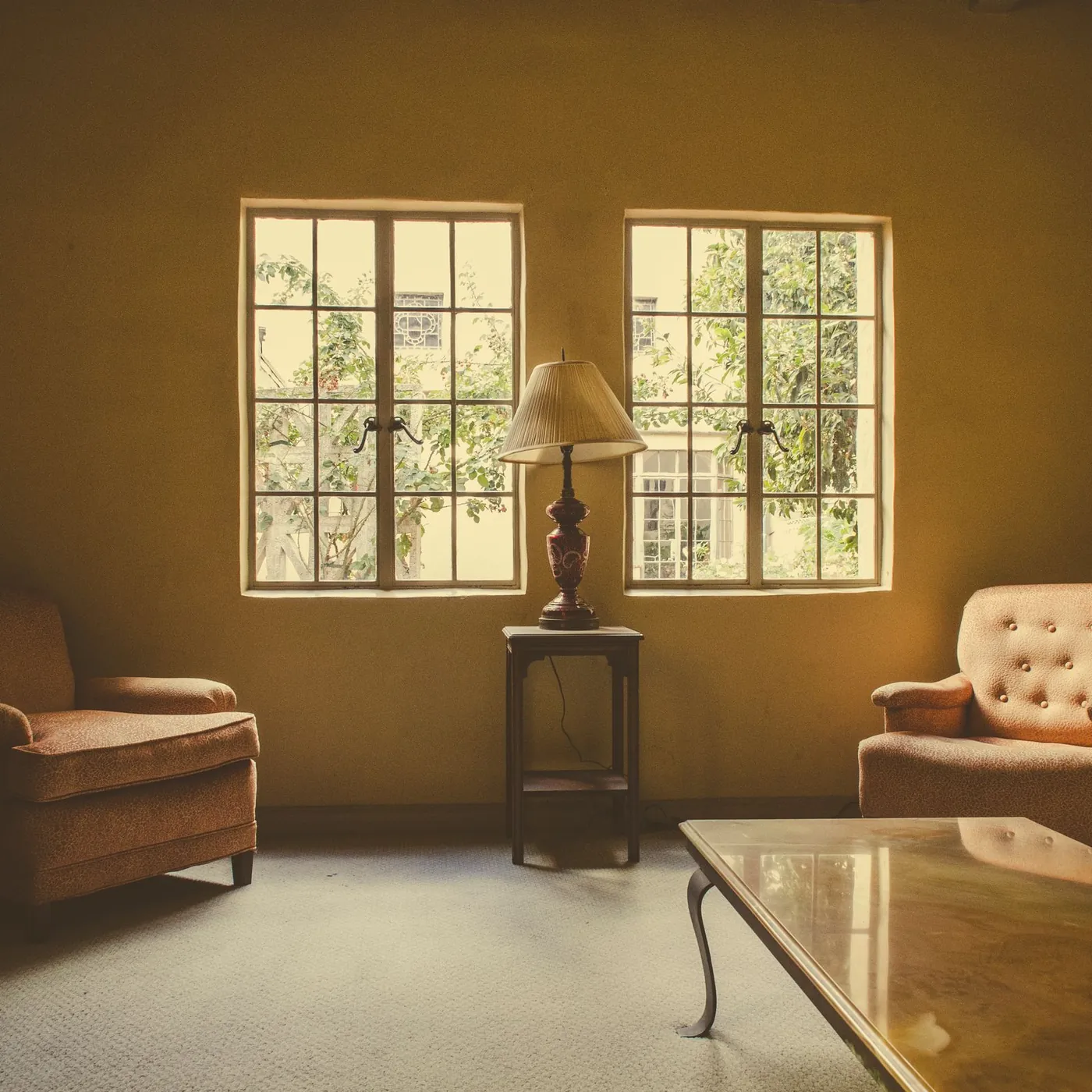 A quiet sitting room in warm amber light: two upholstered armchairs face each other beside a lamp between two garden windows