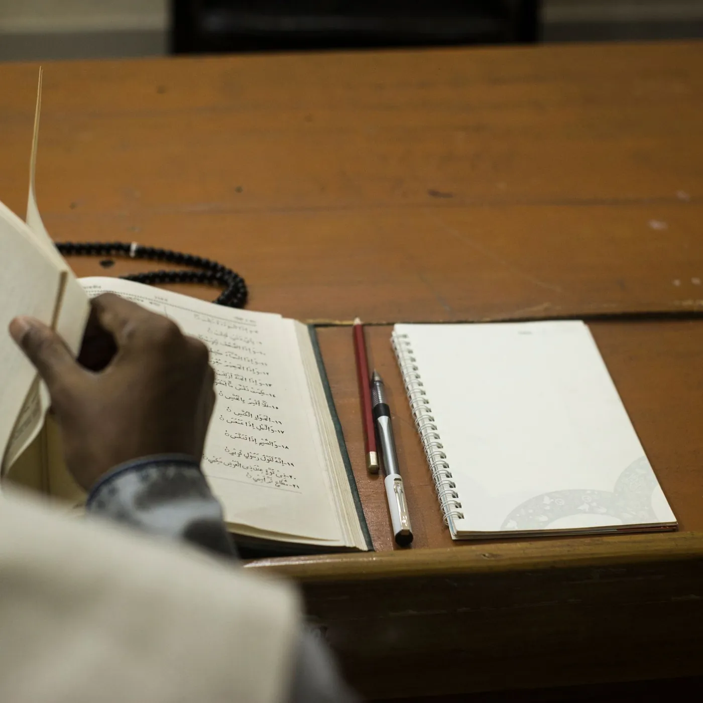 A man's hand turning the pages of an Arabic book beside a spiral notebook, pencil, pen and prayer beads on a wooden desk