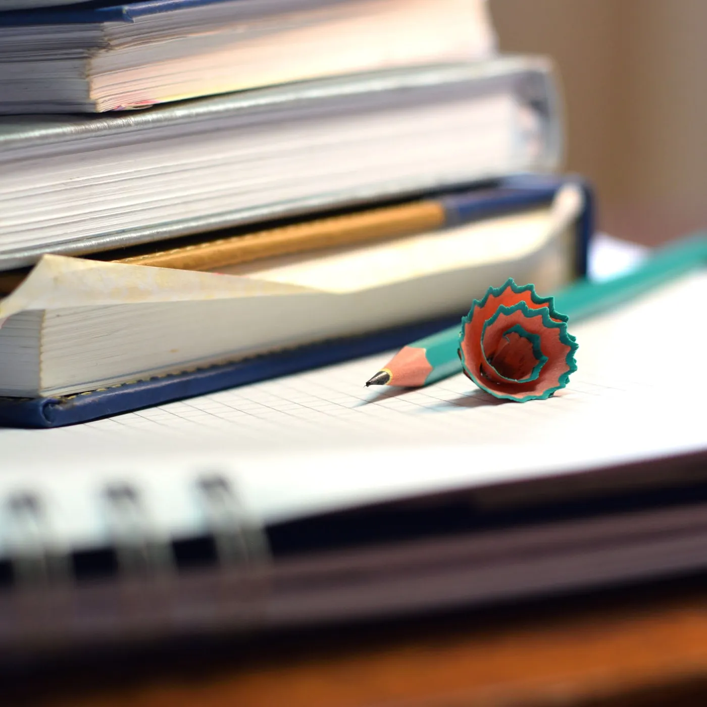 A stack of hardback textbooks over open spiral notebooks, with a freshly sharpened pencil and its curled shaving on top