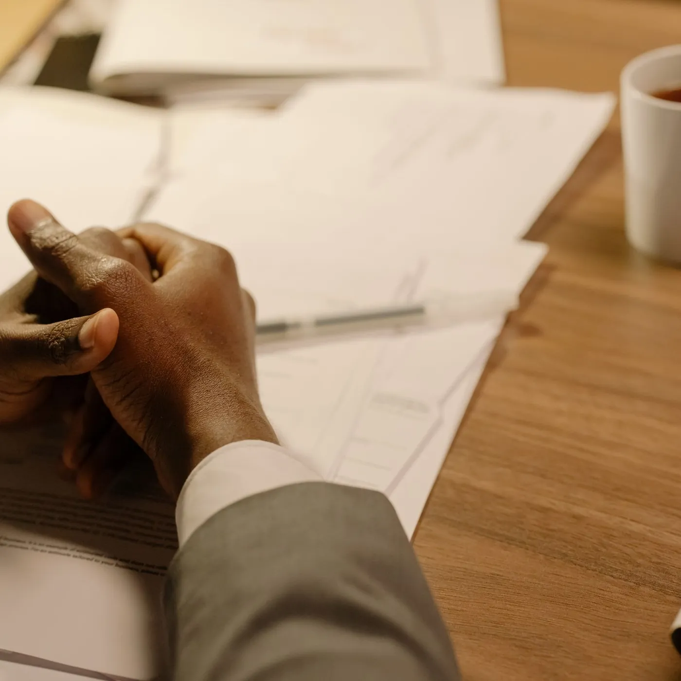 A man's clasped hands resting on papers at a warm wooden table, beside a pen and a white mug of tea in low amber light