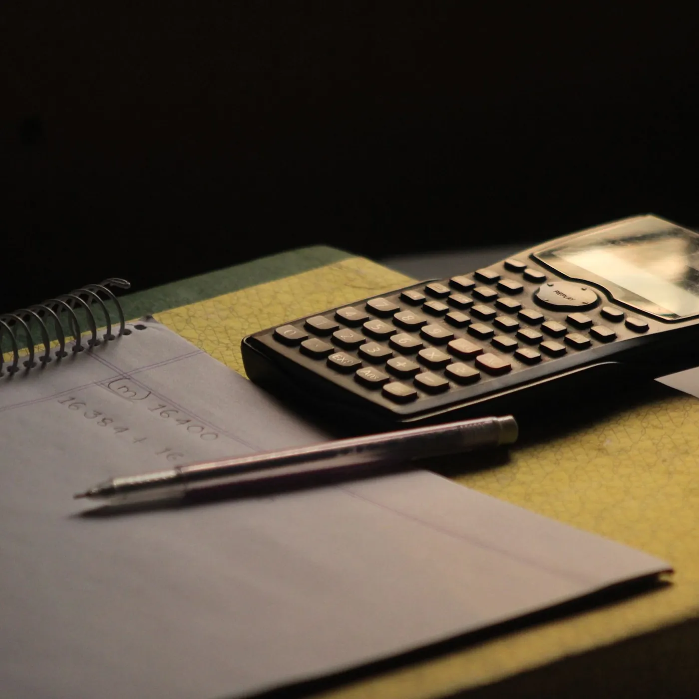 A graphing calculator and a pen resting on an open spiral notebook with pencilled sums, in warm low light